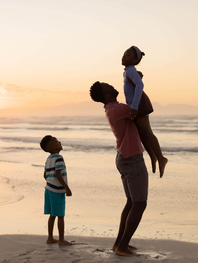 African american son looking at father picking up girl while standing on beach against sky at sunset. nature, unaltered, beach, childhood, family, togetherness, lifestyle, enjoyment and holiday.